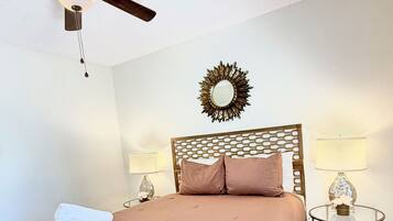 Une chambre à coucher comprenant un lit, un ventilateur de plafond, deux lampes de chevet, un miroir décoratif et une petite table ronde.