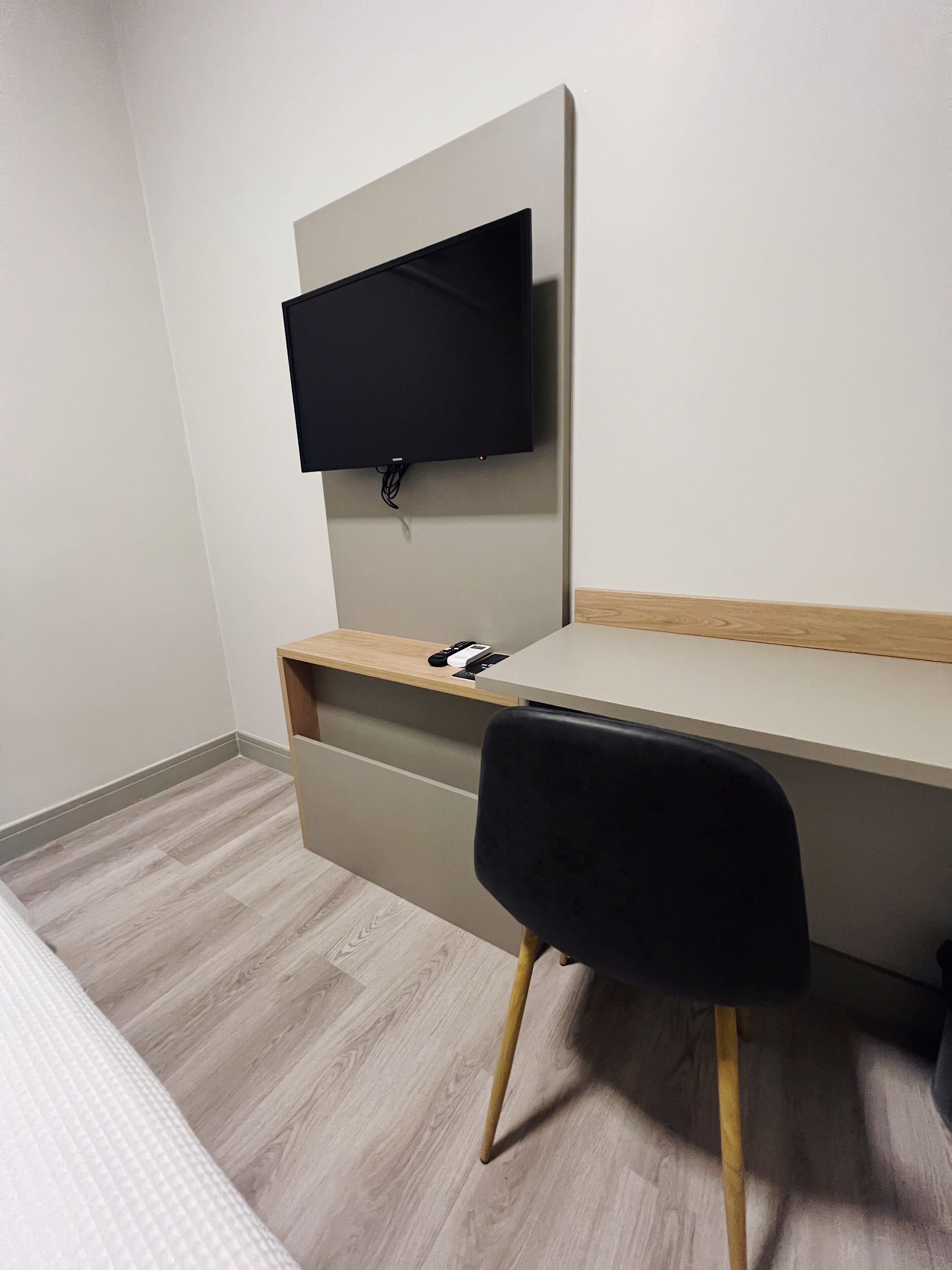 A modern hotel room with a wooden desk, a black chair, and a flat-screen TV mounted on the wall.