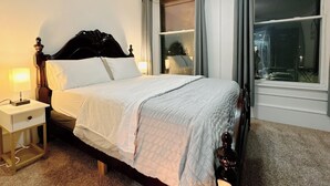 A neatly made bed with a wooden headboard, a bedside table with a lamp, and a window with curtains. - 115#202 / Charming Downtown Dubuque Apartment (Dubuque)
