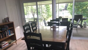 A dining area with a wooden table and chairs, a bookshelf with books, and large windows offering a view of the outdoors.