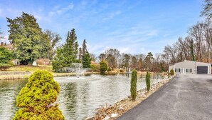 A serene outdoor setting with a pond, a paved walkway, and a building with a garage. - Beautiful Renovated Lake Cottage in SW County and only minutes from Downtown! (Roanoke)
