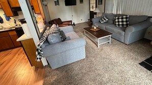 Una sala de estar con un sofá gris, una mesa de centro de madera y un sillón gris. Se ve una cocina al fondo.
