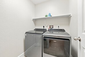 A laundry room with two front-loading washers and dryers, a shelf above the dryers, and a door to the right. - Relaxing Feng Shui Family Retreat 12 Miles from San Antonio Airport (Converse)
