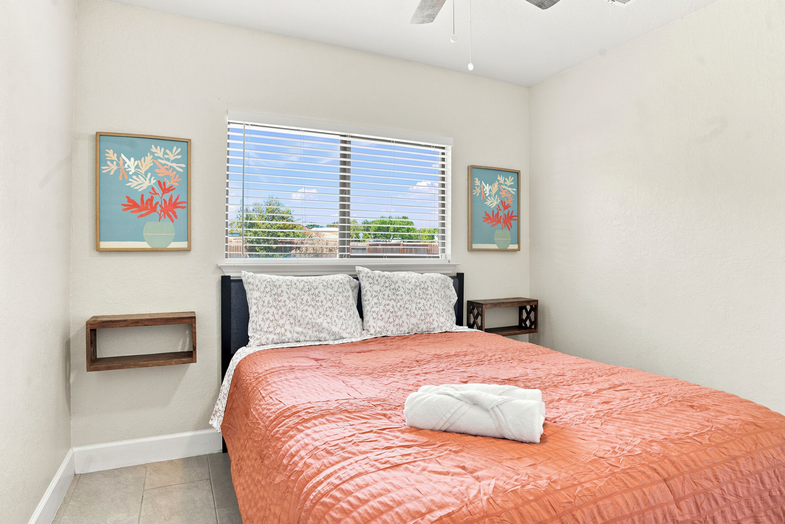 A bedroom with a bed, a window with blinds, two wall-mounted picture frames, a bedside table, and a bedside lamp.