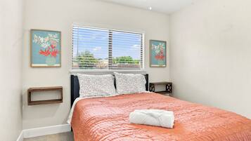 A bedroom with a bed, a window with blinds, two wall-mounted picture frames, a bedside table, and a bedside lamp.