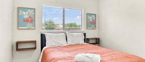 Un dormitorio con cama, ventana con persianas, dos marcos de cuadros en la pared, una mesita de noche y una lámpara de noche.