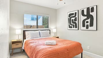 Une chambre à coucher avec un lit, deux lampes de chevet, une fenêtre avec des stores et deux tableaux abstraits accrochés au mur.