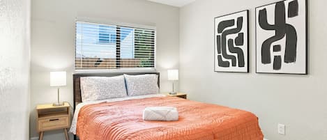 Ein Schlafzimmer mit einem Bett, zwei Nachttischlampen, einem Fenster mit Jalousien und zwei abstrakten Bildern an der Wand.