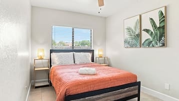 Een slaapkamer met een bed, twee bedlampen, een plafondventilator, een raam met jaloezieën en twee ingelijste foto's aan de muur.