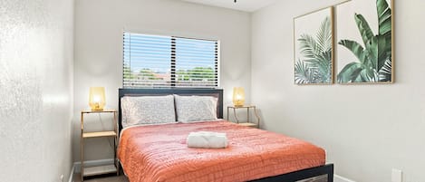A bedroom with a bed, two bedside lamps, a ceiling fan, a window with blinds, and two framed pictures on the wall.