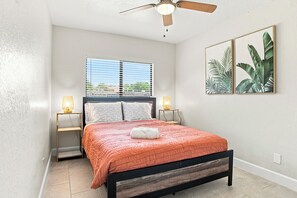 A bedroom with a bed, two bedside lamps, a ceiling fan, a window with blinds, and two framed pictures on the wall.
