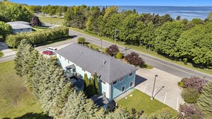 A two-story house with a grey roof, surrounded by lush greenery and a road. - Orchard Beach House Unit #2 (Manistee)