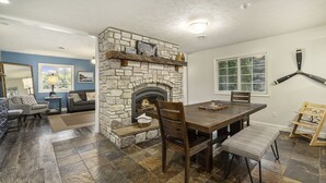 A dining area with a stone fireplace, wooden table, and chairs. - Orchard Beach House Unit #4 (Manistee)