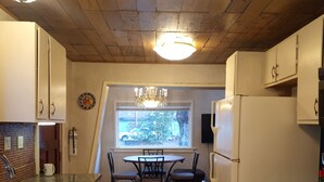 Een keuken met houten plafond, witte kasten, een eettafel met stoelen, een koelkast en een fornuis.