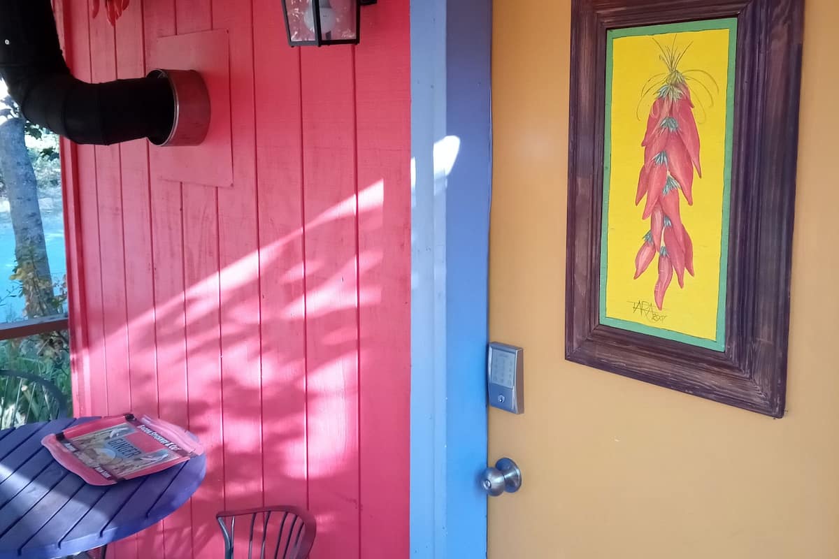 A vibrant room with a red and blue wall, a yellow door, a framed chili pepper artwork, a table with a chair, and a hanging lantern.