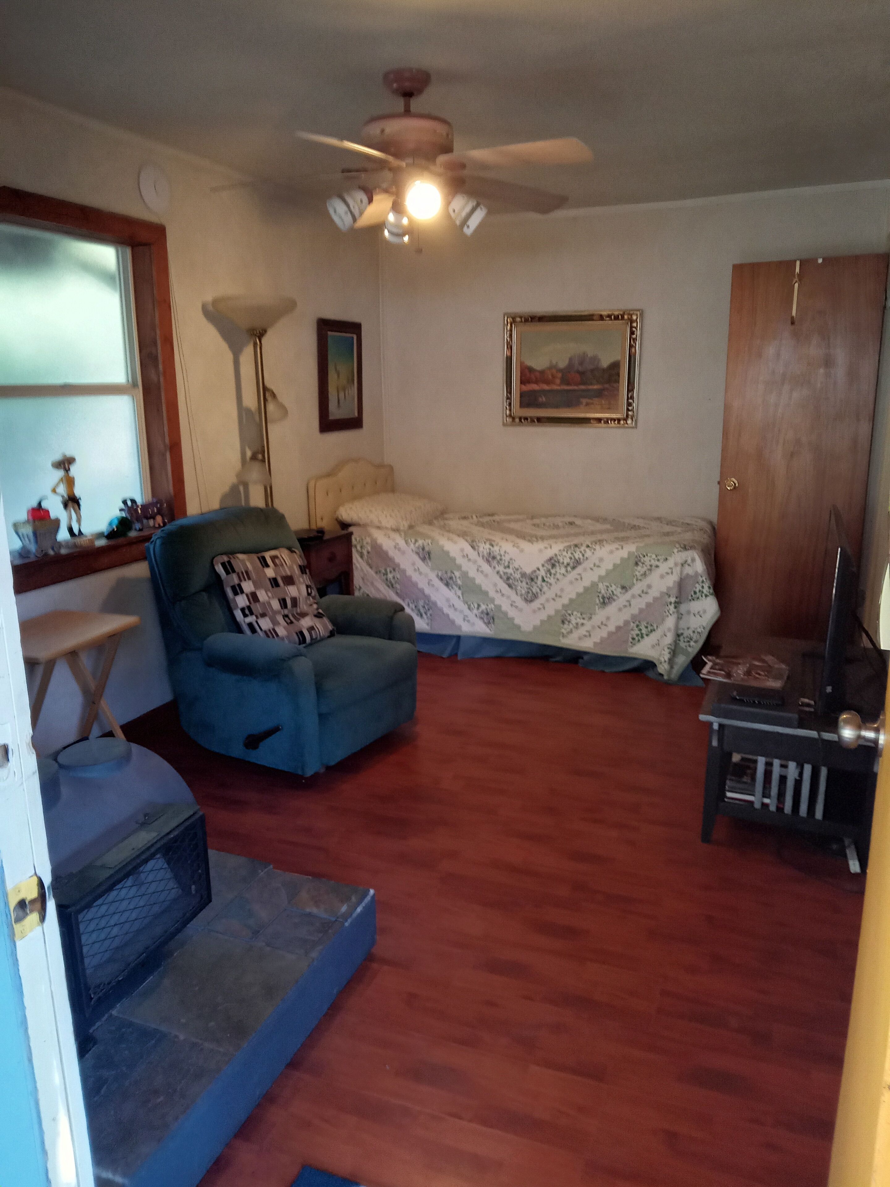 A room with a bed, a blue armchair, a small table, a ceiling fan, a window, and a door.