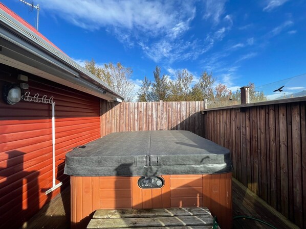 A hot tub in a backyard with a wooden fence, a red building, and a clear blue sky.