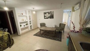 Ett kompakt bostadsutrymme med en säng, en soffa, ett soffbord och en matplats. Det finns inbyggda hyllor och en tavla på väggen.