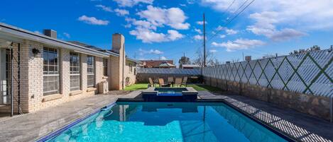 A rectangular swimming pool with a clear blue water surface, surrounded by a brick patio and a metal fence.