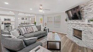 A modern living room with a gray sectional sofa, a flat-screen TV mounted on a stone fireplace, and a ceiling fan. - Beautiful and Spacious, Home - Great location! (San Antonio)