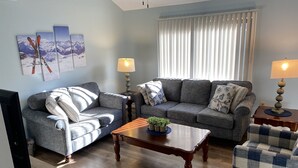 A living room with a grey sofa, a blue armchair, and a wooden coffee table. There are two lamps on side tables and a TV on the left. - Warm and inviting, close to the best New England hiking and outdoor fun! (Bartlett)