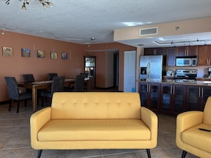 A modern living room with a yellow sofa, dining area with chairs, and a kitchen with stainless steel appliances.