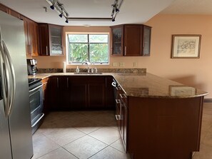 A kitchen with a stainless steel refrigerator, a built-in oven, a sink with a faucet, and wooden cabinets. There is a window with a view of greenery outside.