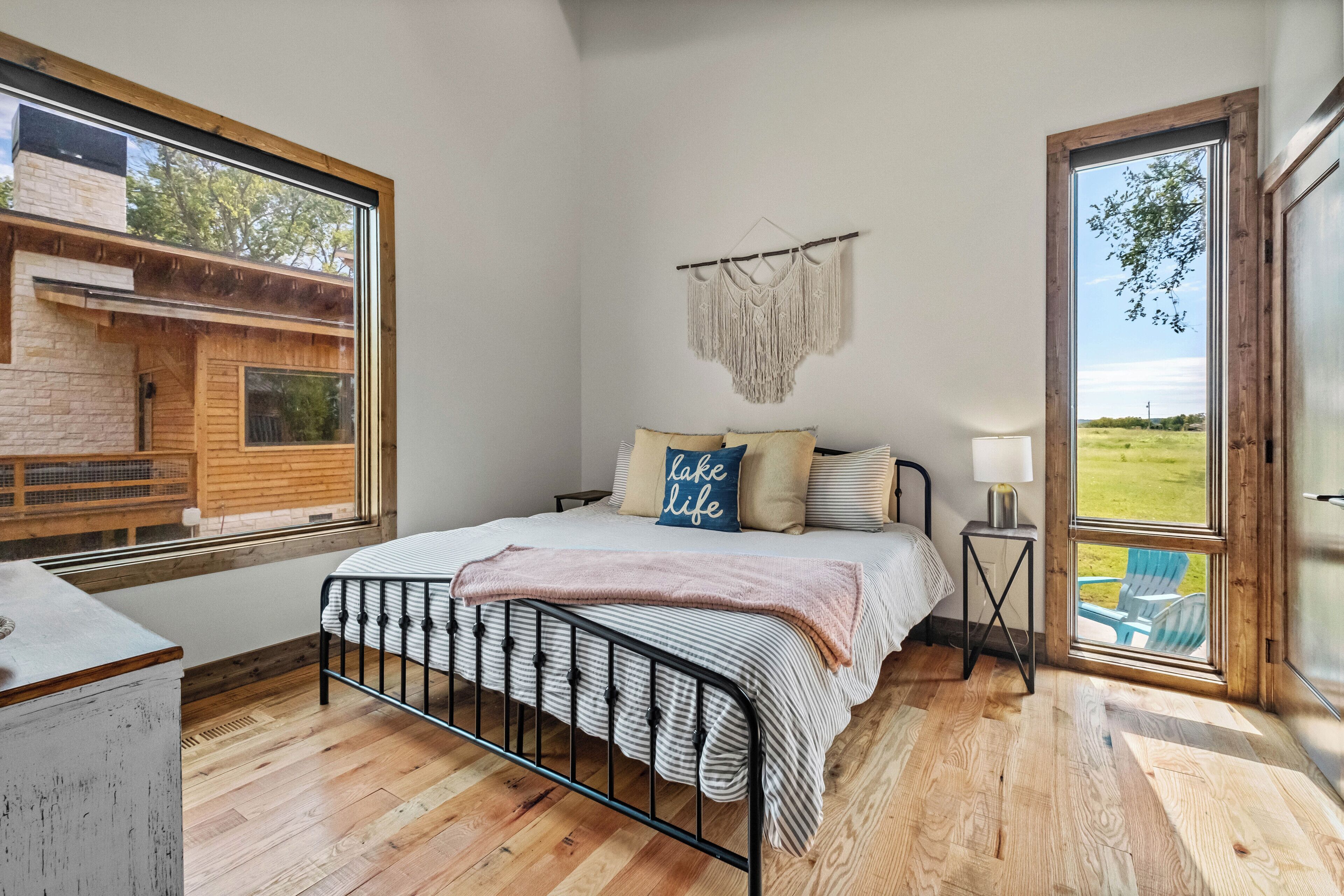 Una camera da letto con un'ampia finestra, un letto con testiera, un arazzo macramè e vista su una casa e del verde all'esterno.