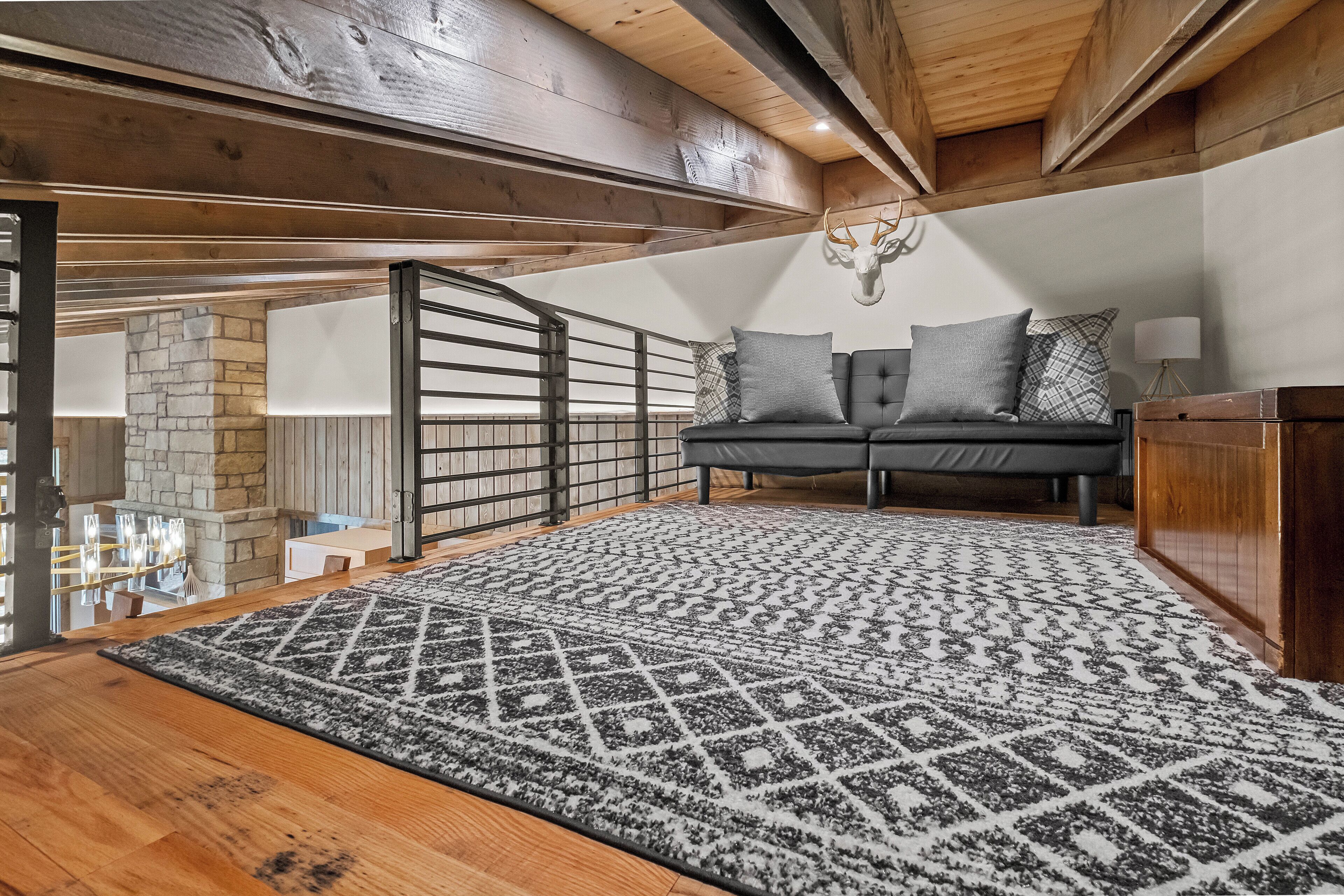 Quarto tipo loft acolhedor, com teto de madeira, parede de pedra, sofá preto com almofadas estampadas e um tapete grande.
