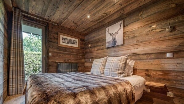 A wooden cabin bedroom with a bed, a window with curtains, a radiator, and two framed pictures on the wall.