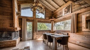 A rustic cabin interior with a wooden dining table, chairs, a fireplace, and large windows offering views of the outdoors.