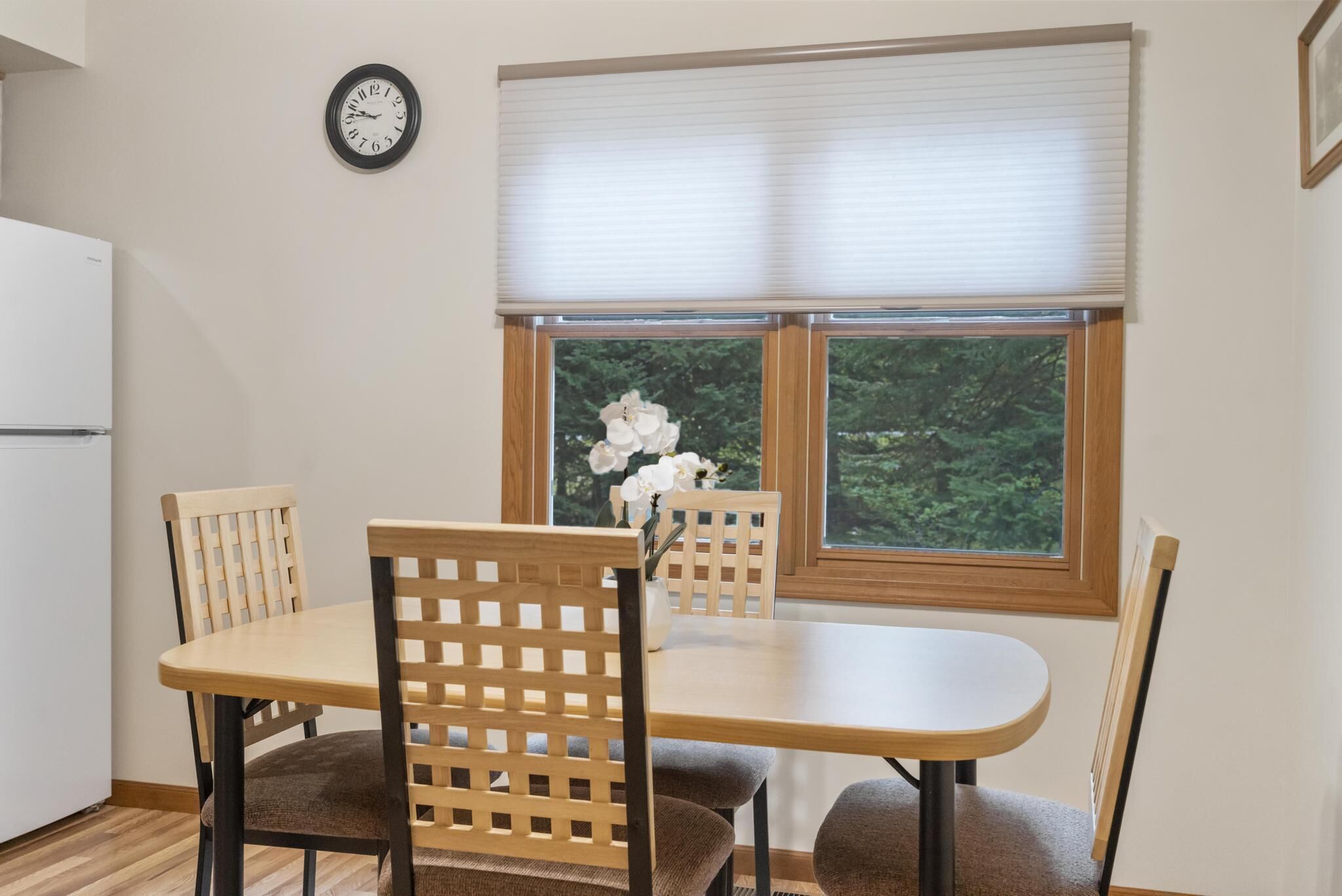 Un coin repas avec une table et des chaises, une horloge au mur et une fenêtre donnant sur un paysage verdoyant.