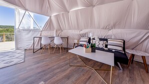 Una habitación con carpa, suelo de madera, una mesa blanca, dos sillas blancas, un sofá con cojines estampados y vistas al exterior con vegetación.