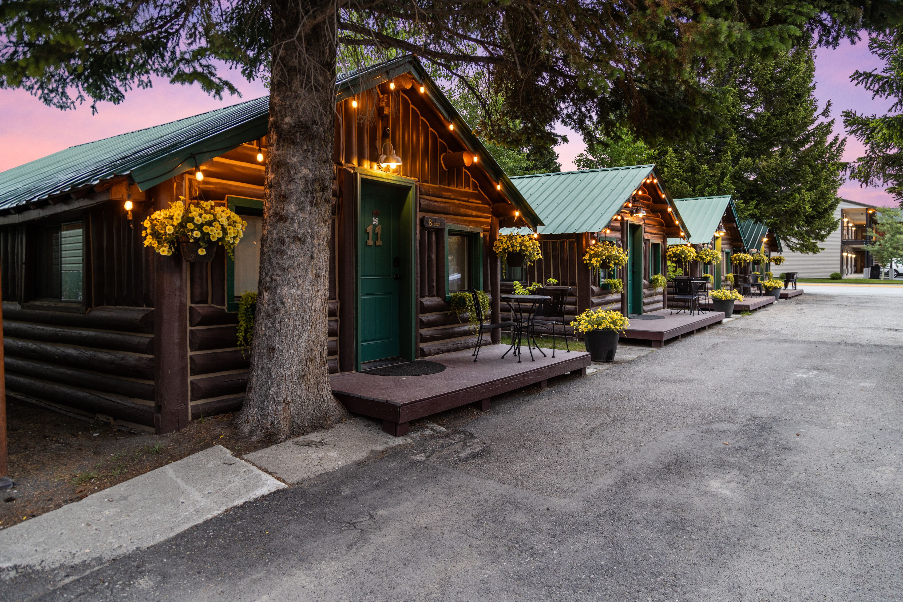 A row of log cabins with green roofs and hanging flower baskets, each with a porch and outdoor seating.
