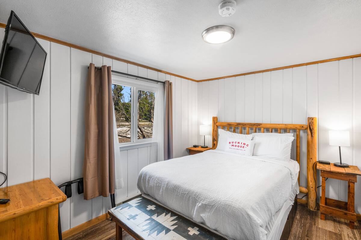 Un dormitorio con cama de madera, televisor montado en la pared, mesita de noche con lámpara y ventana con cortinas.