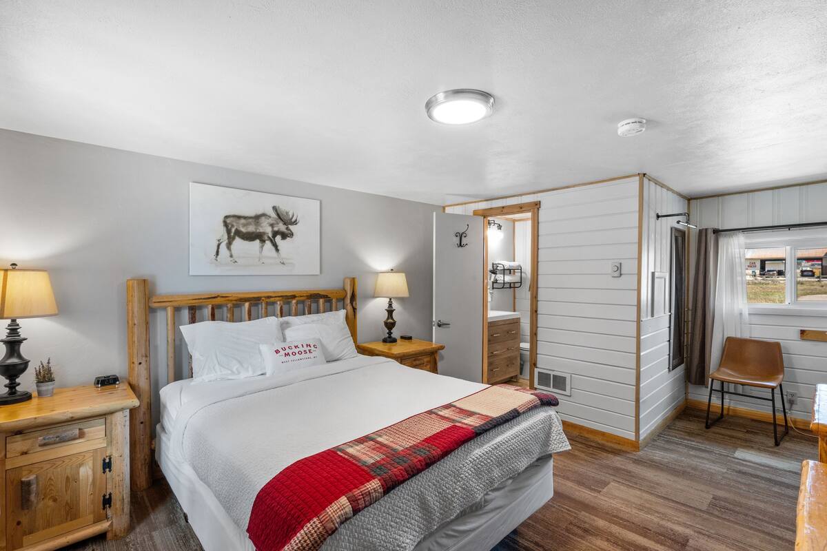 Un dormitorio con una cama de madera, un edredón rojo y gris, un cuadro de un alce en la pared y una mesita de noche de madera con una lámpara.