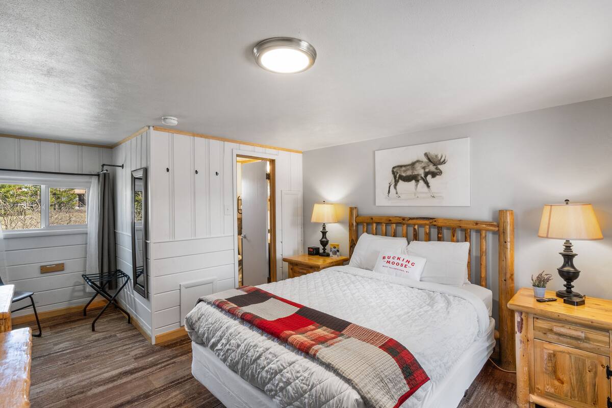 Un dormitorio con una cama grande, cabecera de madera, mesita de noche con lámpara, ventana con cortinas y una puerta que da a otra habitación.