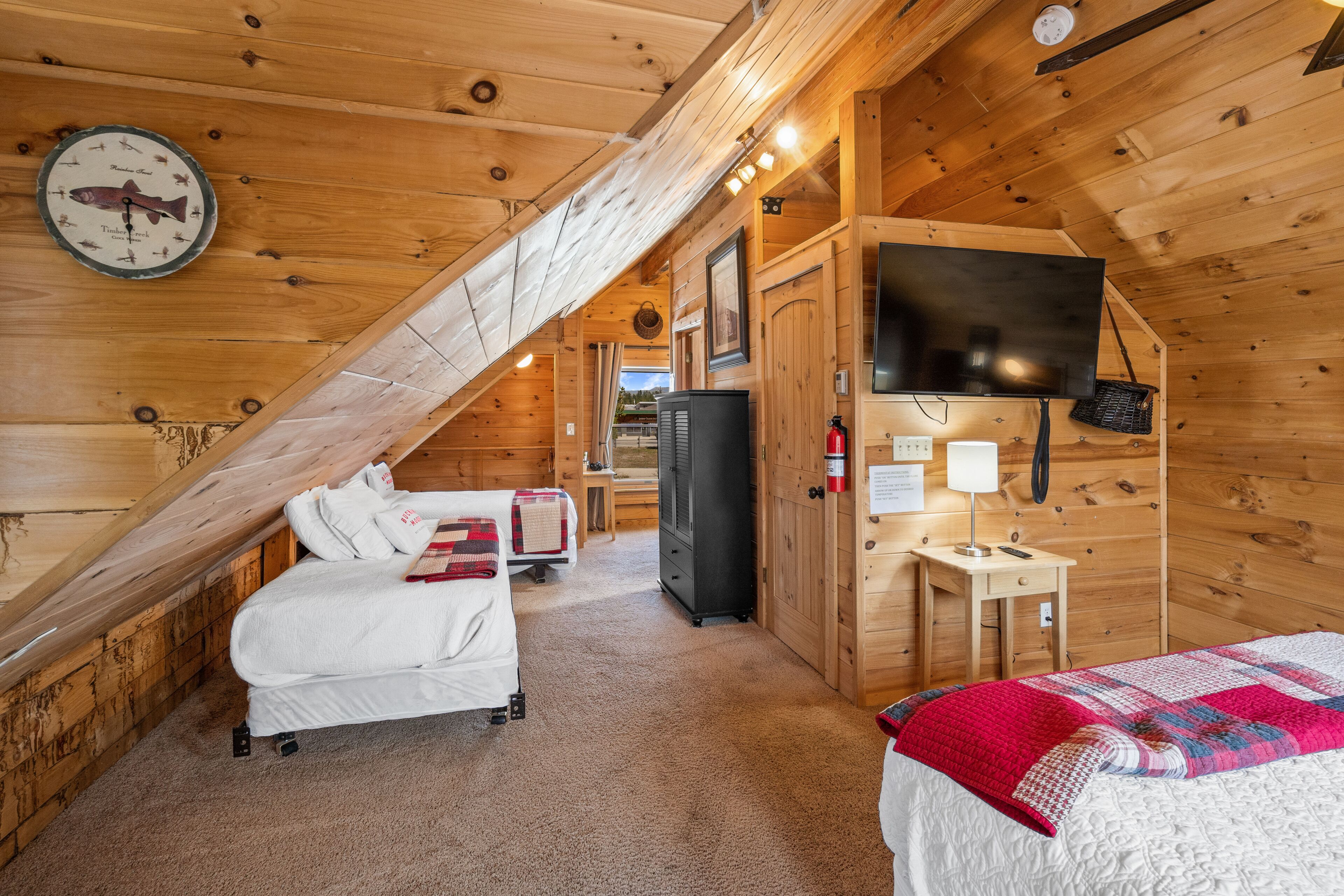Una stanza accogliente di una baita in legno, con un letto, una scrivania, un televisore e un orologio appeso al muro.