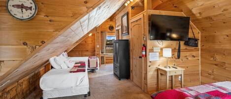 Una stanza accogliente di una baita in legno, con un letto, una scrivania, un televisore e un orologio appeso al muro.