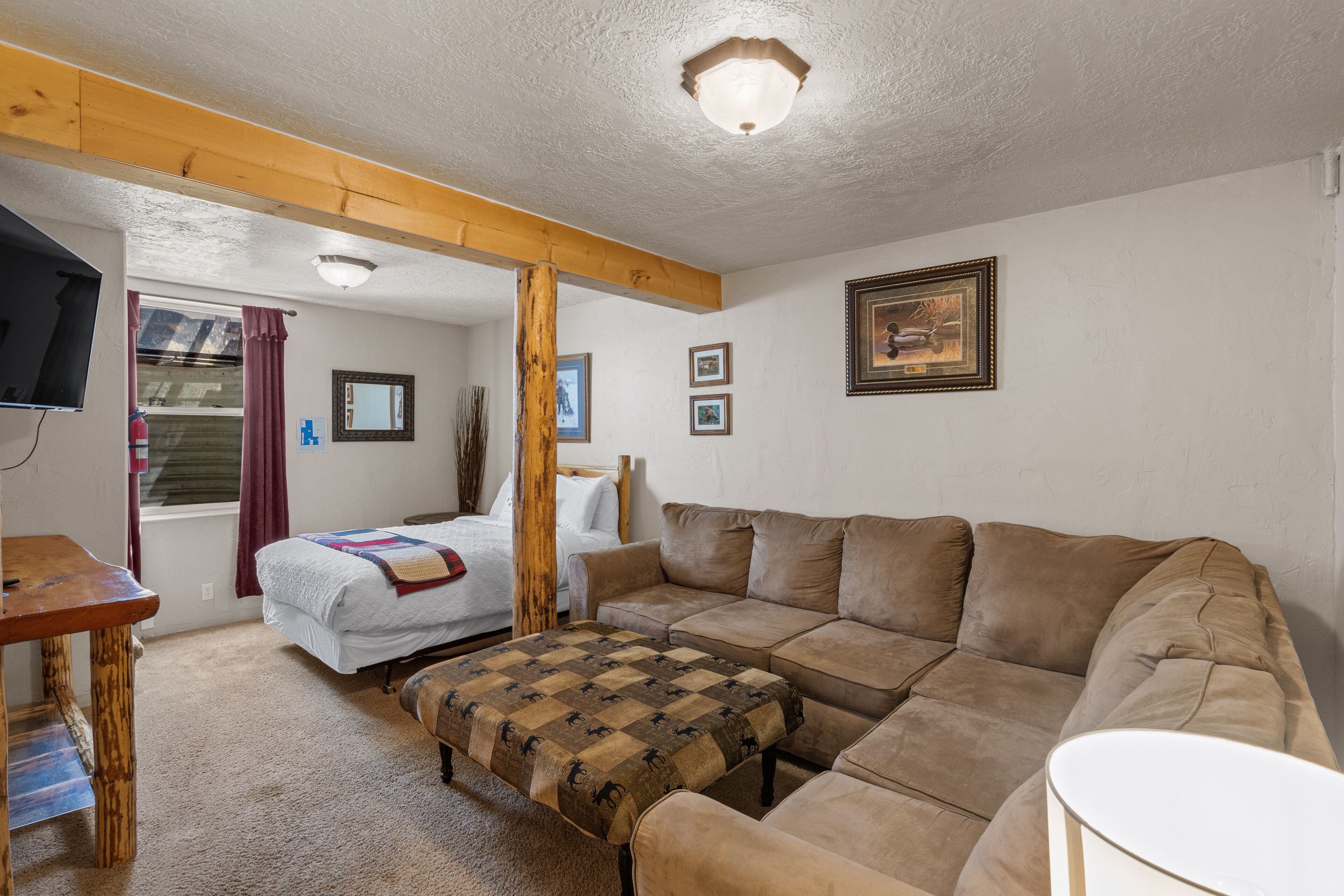 A cozy room with a wooden ceiling, a large brown sofa, a small table, a bed with a quilt, and a framed picture on the wall.