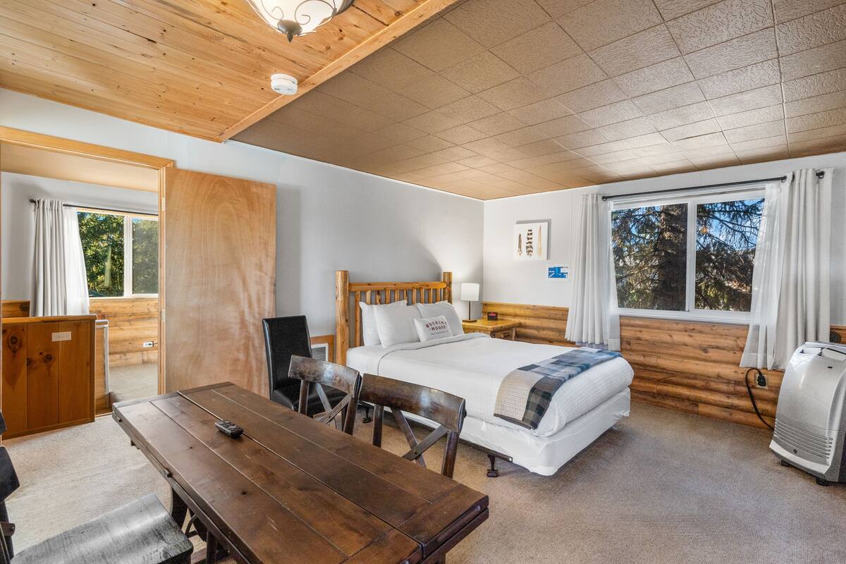 Una habitación tipo cabaña de madera con una cama, una mesa de madera, una silla, un escritorio, un calentador y una ventana con cortinas.