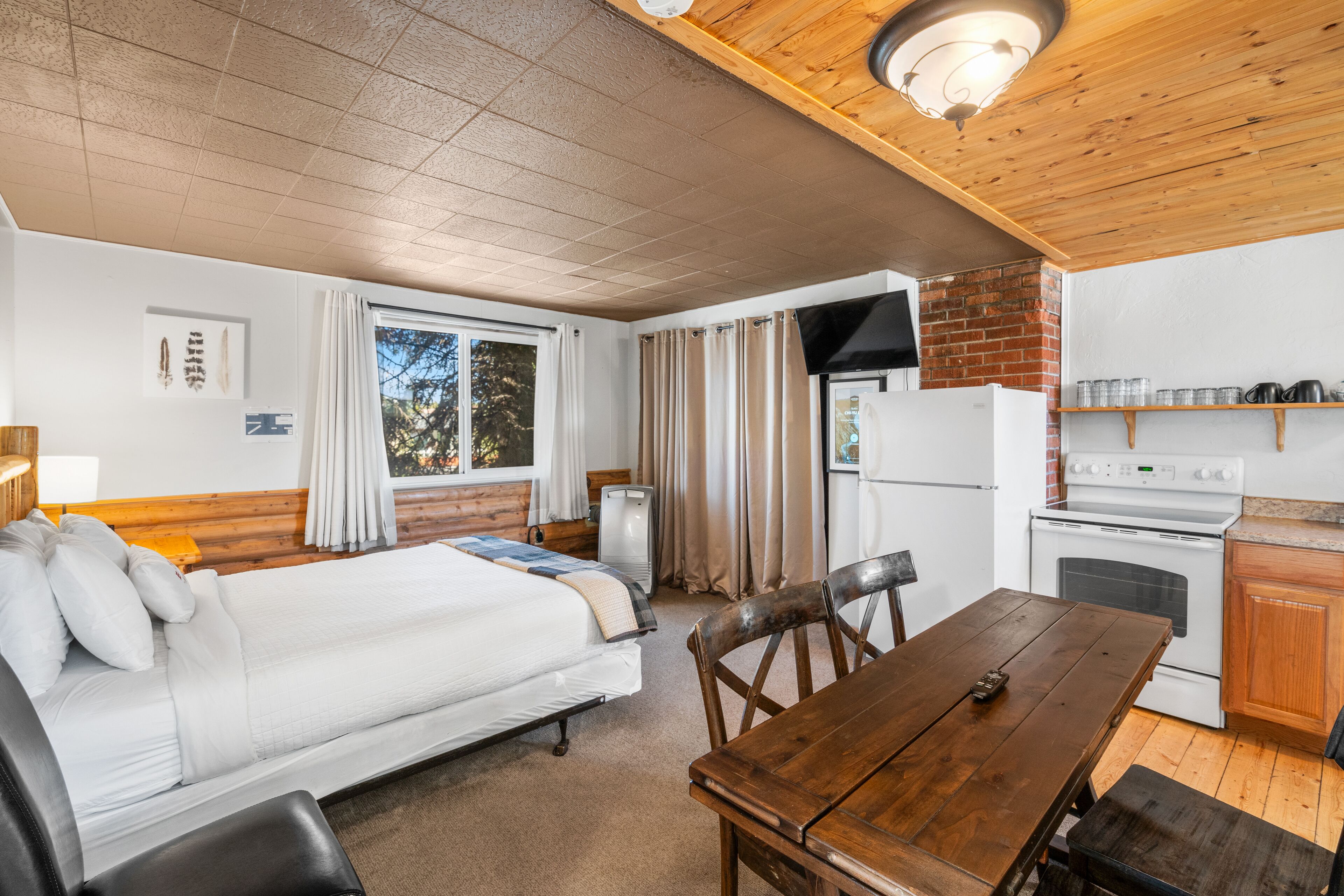 A compact room with a bed, a wooden dining table, a kitchen area with a stove and refrigerator, and a window with curtains.