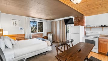 A compact room with a bed, a wooden dining table, a kitchen area with a stove and refrigerator, and a window with curtains.