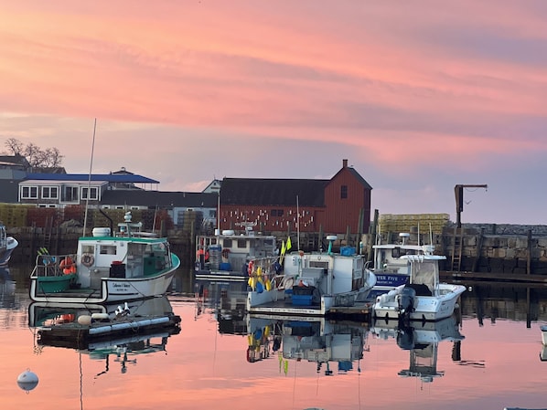 A marina with boats docked, a red building, and a calm water surface reflecting the surroundings.