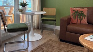 A living room with a view of a building through large windows, a brown sofa with a palm tree pillow, a wicker chair, a white table with a plant, and a green wall. - Newly Remodeled. Water views from your private lanai.  Steps to Madeira Beach. (Madeira Beach)