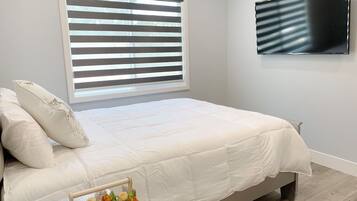 Un dormitorio con cama, ventana con persianas, televisor de pantalla plana montado en la pared y una mesita de noche con un objeto decorativo.