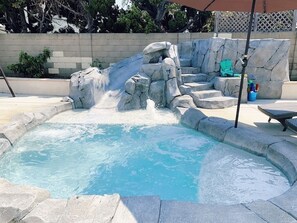A swimming pool with a rock waterfall feature, surrounded by stone steps and a lounge area with a table and chairs.