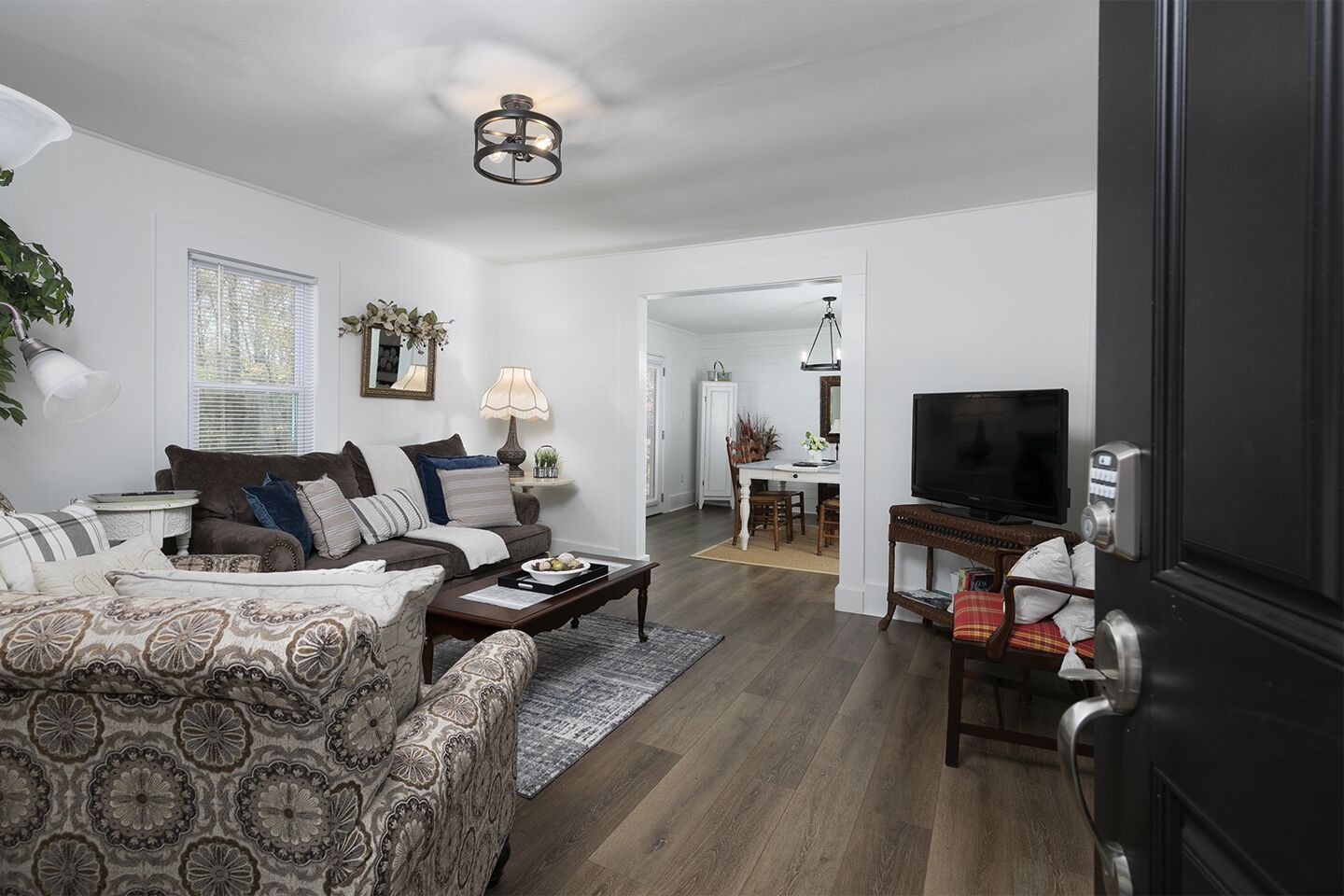A living room with a sofa, coffee table, and TV stand. There is a dining area with a table and chairs. The room has wooden flooring and white walls.