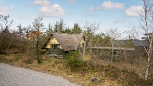 En liten, rustik stuga med ett sluttande tak, omgiven av träd och en grusgång.