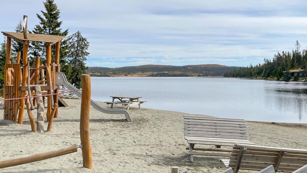 A wooden play structure with a slide and climbing area, a picnic table, and a bench on a sandy beach with a lake and hills in the background. - Stunning home in Sjusjøen with sauna (Sjusjøen)